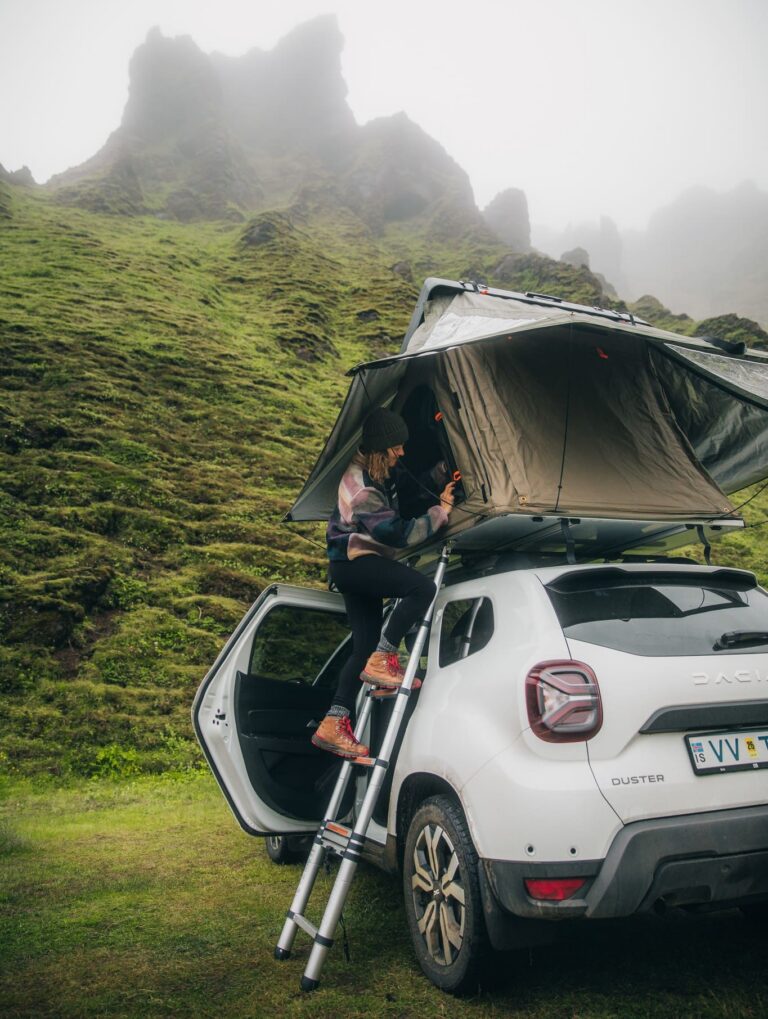 person climbing into a rooftop tent on a car in a foggy, green mountain landscape